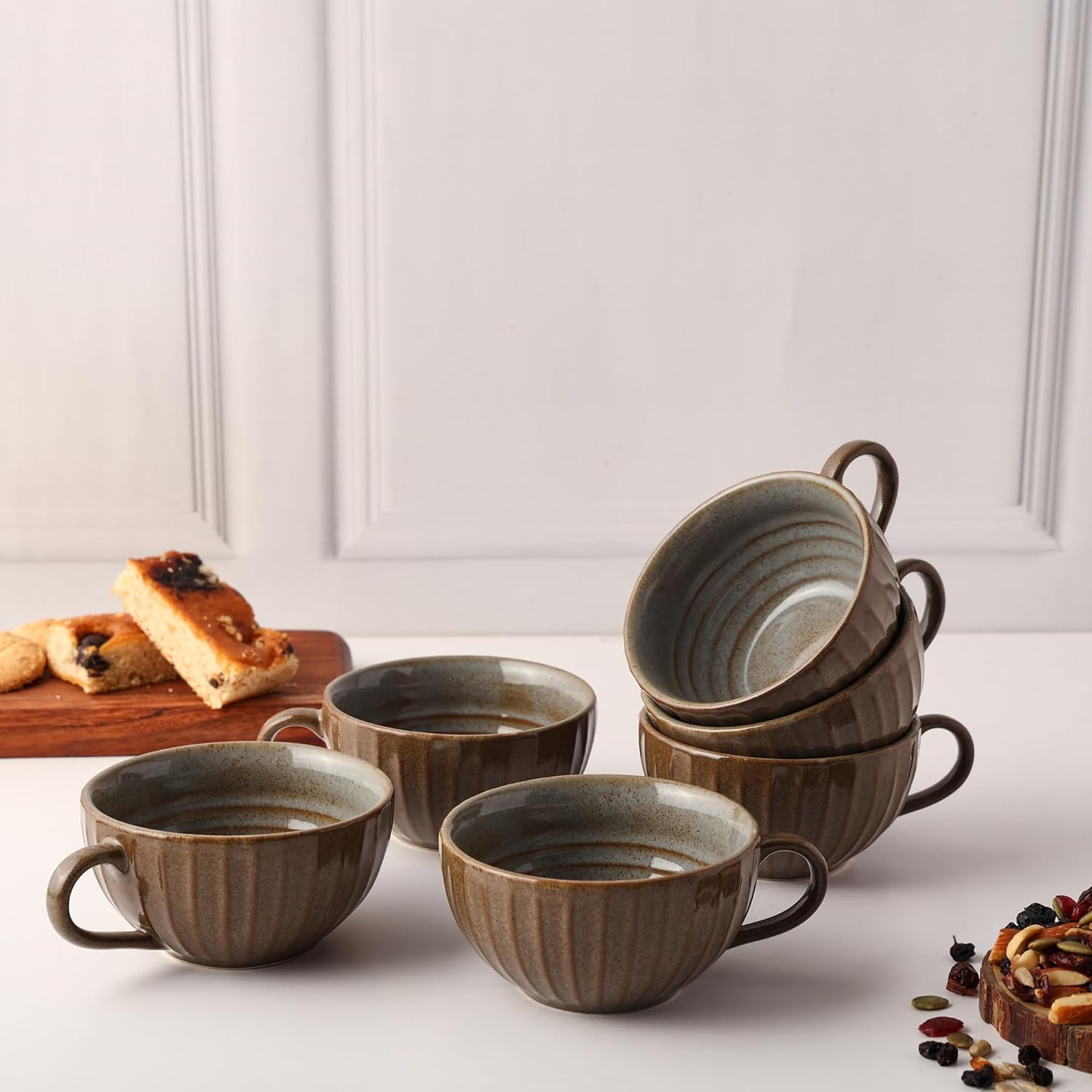Set of ceramic mugs on a white surface with a wooden board of bread and nuts.
